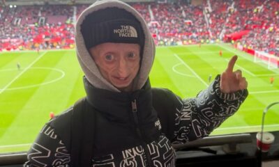 Pessoa com progeria sorri, vestindo The North Face, apontando, em estádio de futebol
