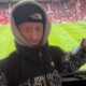 Pessoa com progeria sorri, vestindo The North Face, apontando, em estádio de futebol