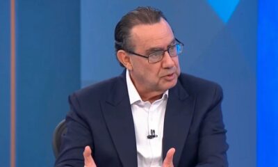 Homem de óculos, terno escuro, camisa branca, gesticula falando em estúdio de TV
