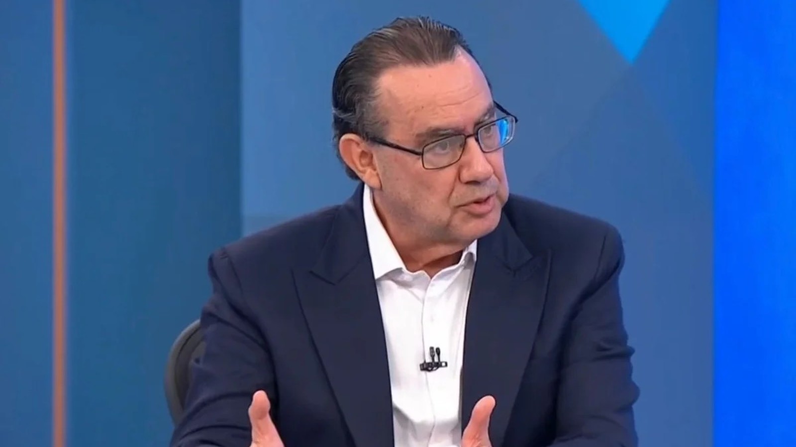 Homem de óculos, terno escuro, camisa branca, gesticula falando em estúdio de TV