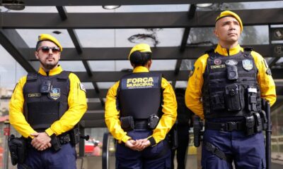 Três guardas municipais em uniforme amarelo e azul com coletes pretos e boinas amarelas em formação