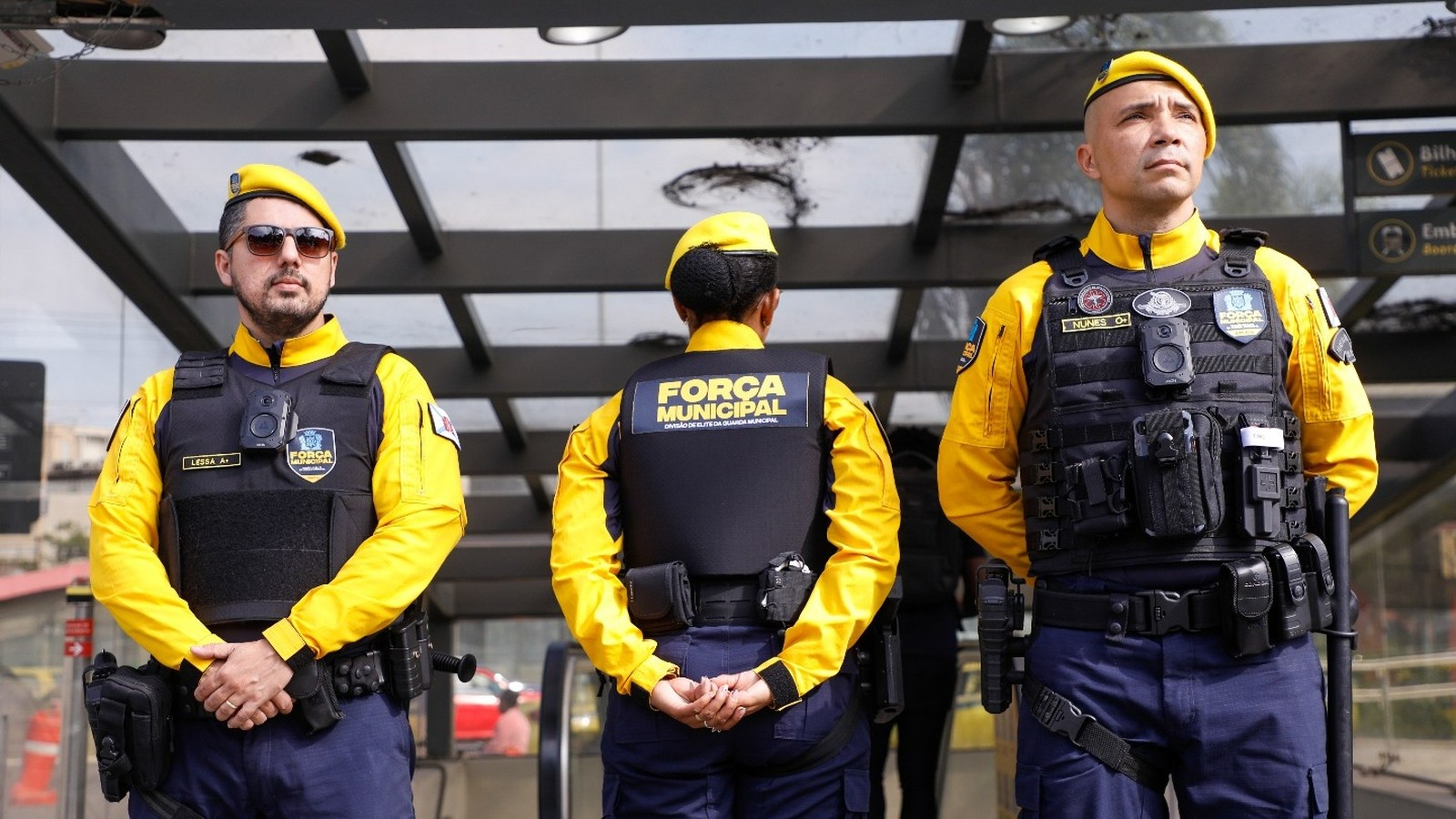 Três guardas municipais em uniforme amarelo e azul com coletes pretos e boinas amarelas em formação