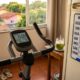 Bicicleta ergométrica em ambiente doméstico com painel marcando 15 minutos e calendário indicando quatro sessões semanais.