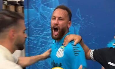 Jogador Neymar com boca aberta, camisa azul de futebol, braços de duas pessoas