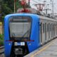 Trem azul e cinza da SuperVia na plataforma, dois homens caminham, com vegetação e trilhos