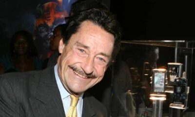 Homem sorridente de terno com bigode em evento, ao fundo expositor de produtos Transformers