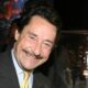 Homem sorridente de terno com bigode em evento, ao fundo expositor de produtos Transformers