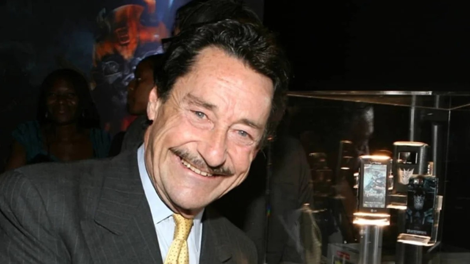 Homem sorridente de terno com bigode em evento, ao fundo expositor de produtos Transformers