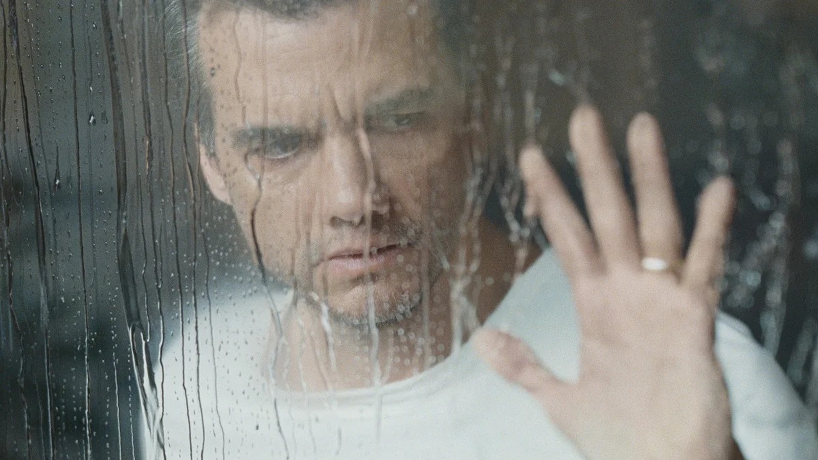 Homem sério através de vidro com chuva, sua mão com anel toca a superfície