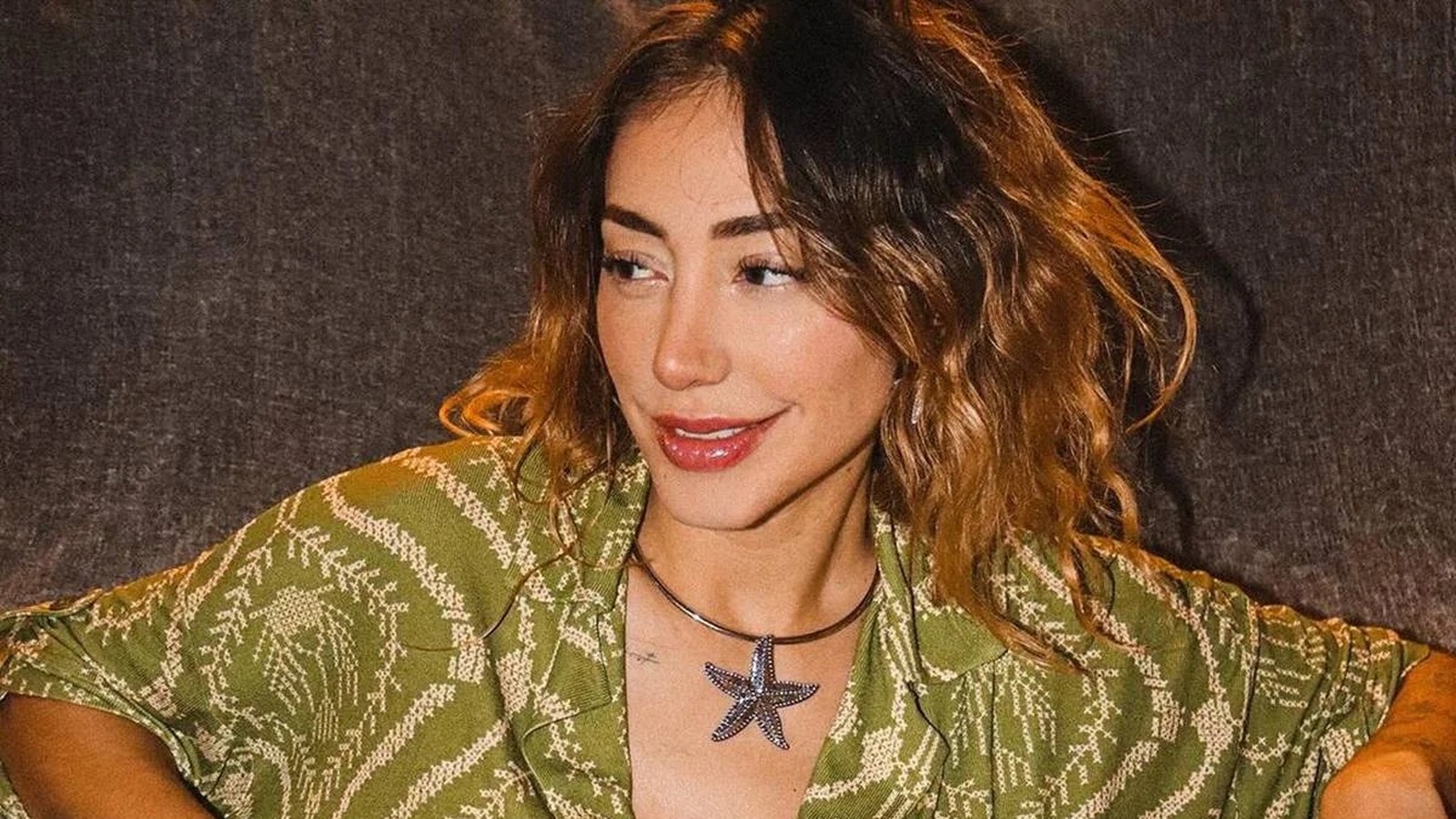 Mulher com cabelo castanho ondulado e camisa verde estampada usando colar com estrela-do-mar