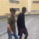 Policial escolta homem de camisa preta na rua, com viatura e prédio amarelo ao fundo