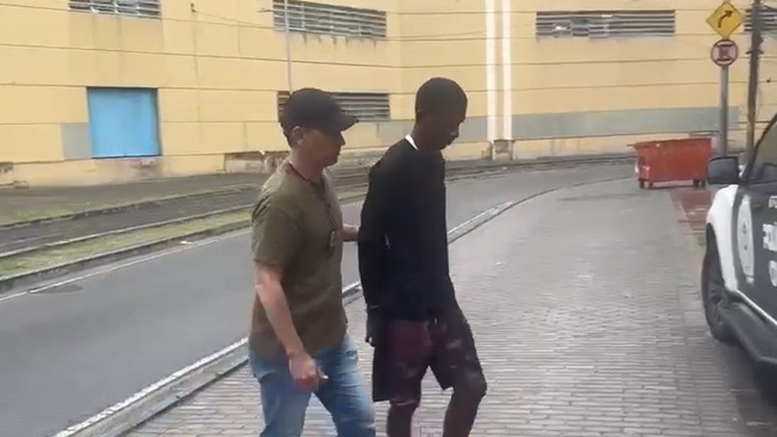 Policial escolta homem de camisa preta na rua, com viatura e prédio amarelo ao fundo