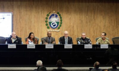 Sete autoridades, incluindo militares, sentadas em uma mesa de conferência com placas de identificação e brasão