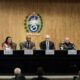 Sete autoridades, incluindo militares, sentadas em uma mesa de conferência com placas de identificação e brasão