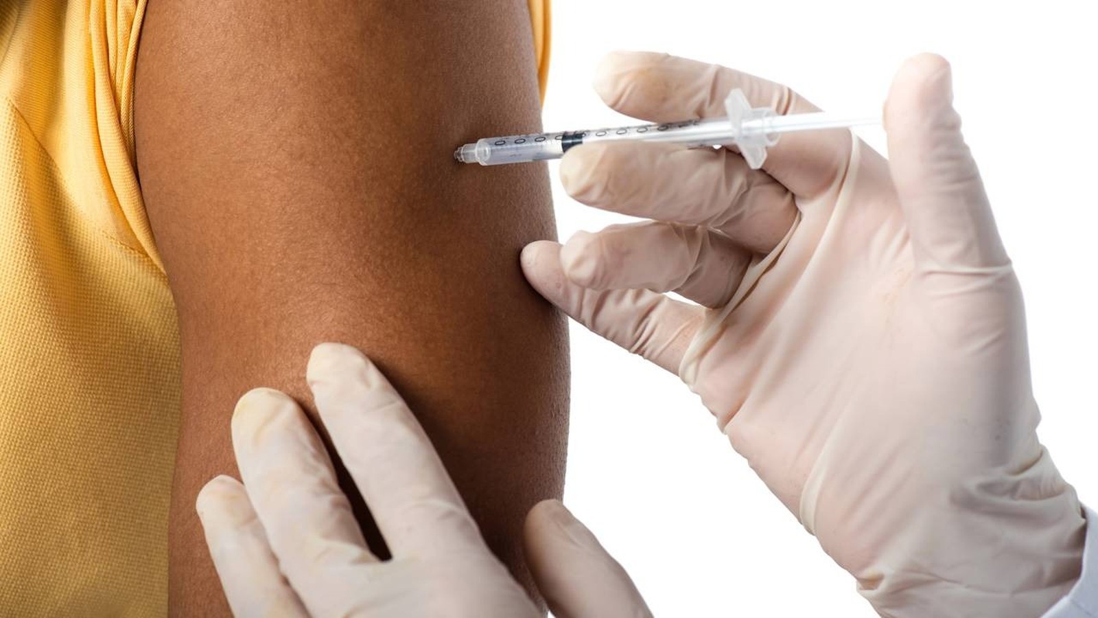 Mão enluvada aplicando injeção com seringa no braço de pessoa em camisa amarela