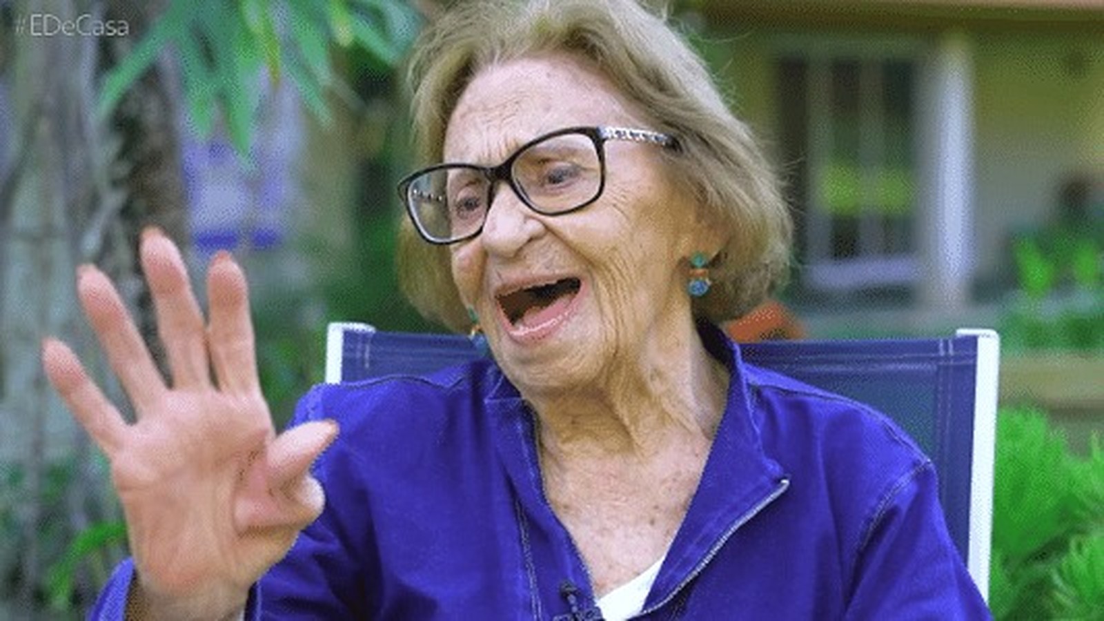 Laura Cardoso de óculos, boca aberta, gesticula mão. Logotipo #EDeCasa