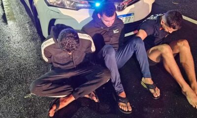 Três homens sentados no chão molhado, próximo ao para-choque de carro branco com luz azul