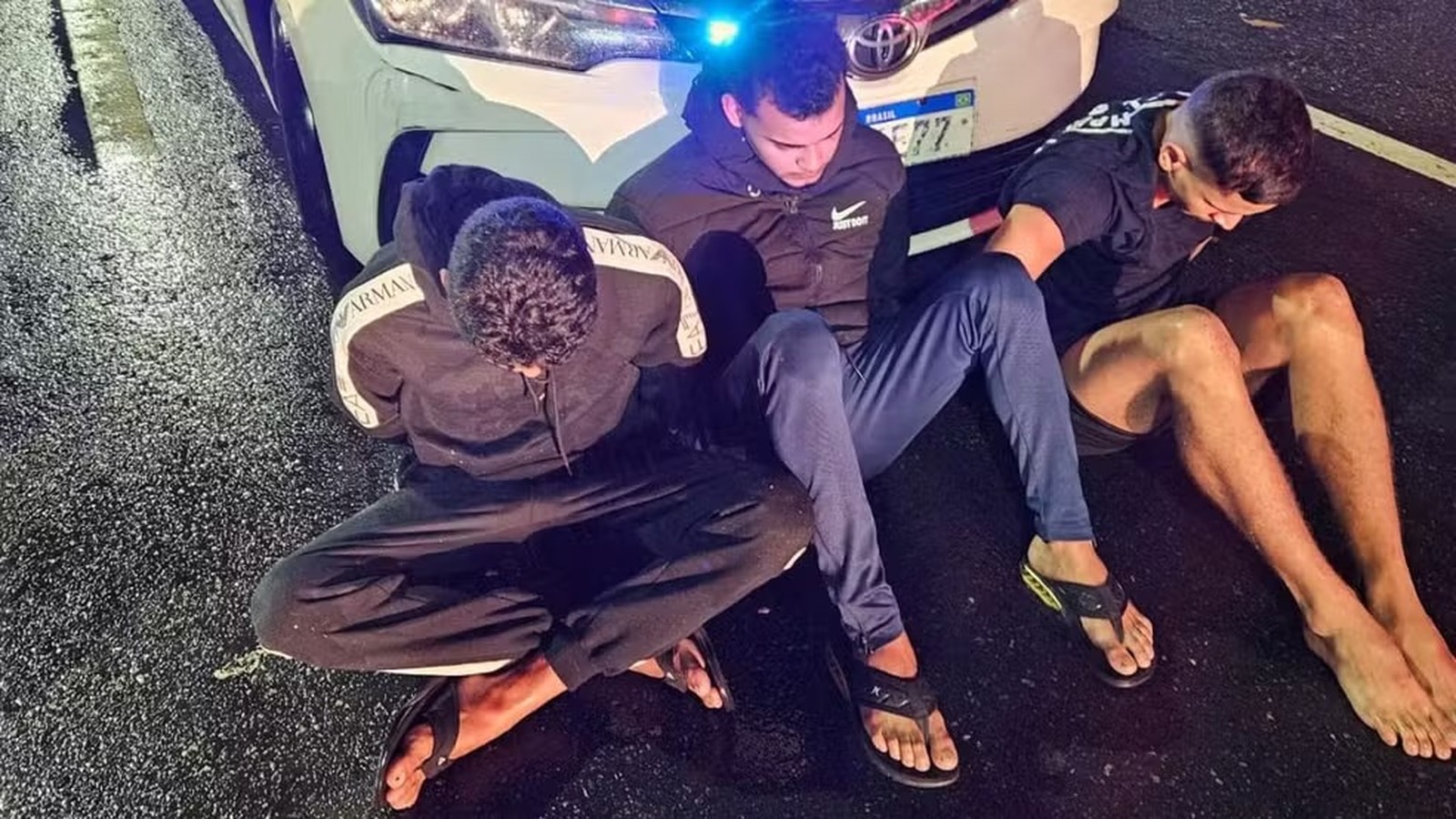 Três homens sentados no chão molhado, próximo ao para-choque de carro branco com luz azul