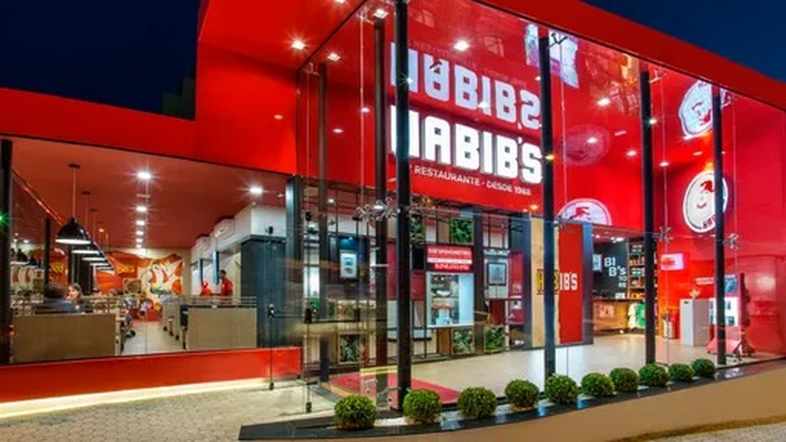 Restaurante Habib's com fachada vermelha, nome em branco, janelas de vidro, interior iluminado, pessoas