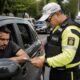 Agente de trânsito com colete amarelo entrega papéis a motorista em carro preto.