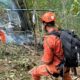 Três socorristas equipados em uniformes laranja operam perto de destroços de helicóptero fumegante em mata fechada