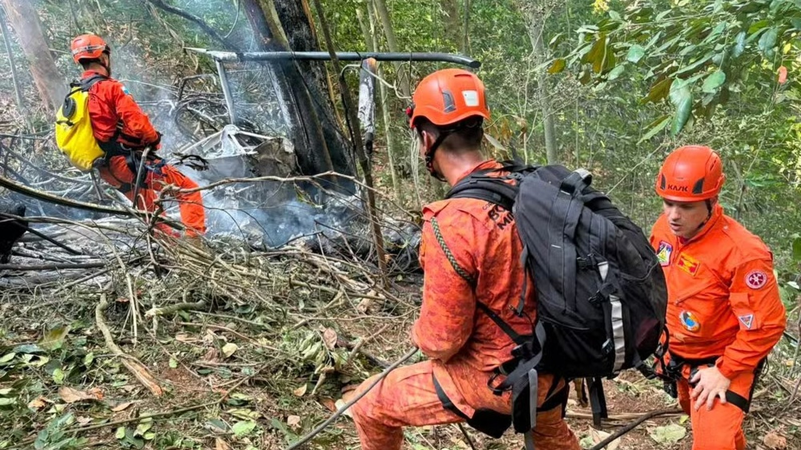 Três socorristas equipados em uniformes laranja operam perto de destroços de helicóptero fumegante em mata fechada