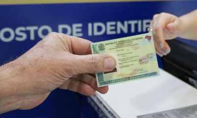 Mão masculina entrega Carteira de Identidade Nacional para mão feminina, em posto de identificação