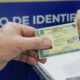 Mão masculina entrega Carteira de Identidade Nacional para mão feminina, em posto de identificação