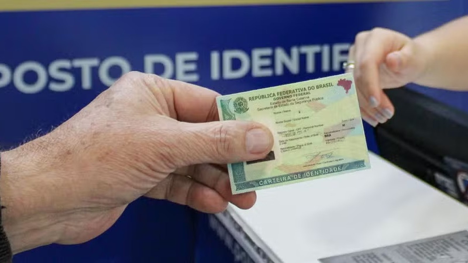 Mão masculina entrega Carteira de Identidade Nacional para mão feminina, em posto de identificação