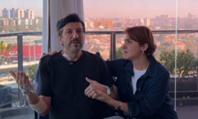 Homem e mulher em varanda, ele com aparelho no pescoço, de mãos dadas, com vista urbana.