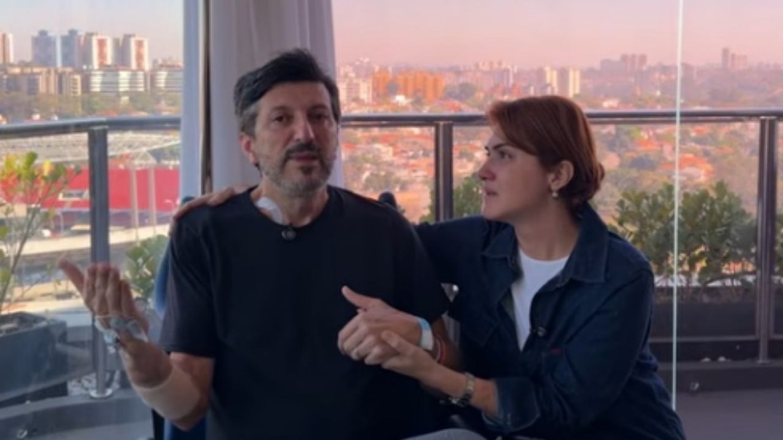 Homem e mulher em varanda, ele com aparelho no pescoço, de mãos dadas, com vista urbana.