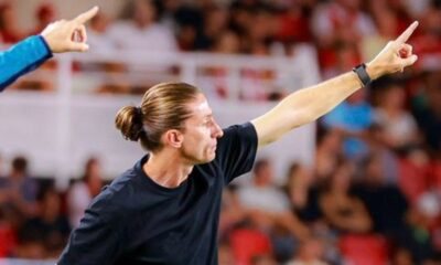 Homem com coque e camiseta preta aponta, em estádio com público ao fundo