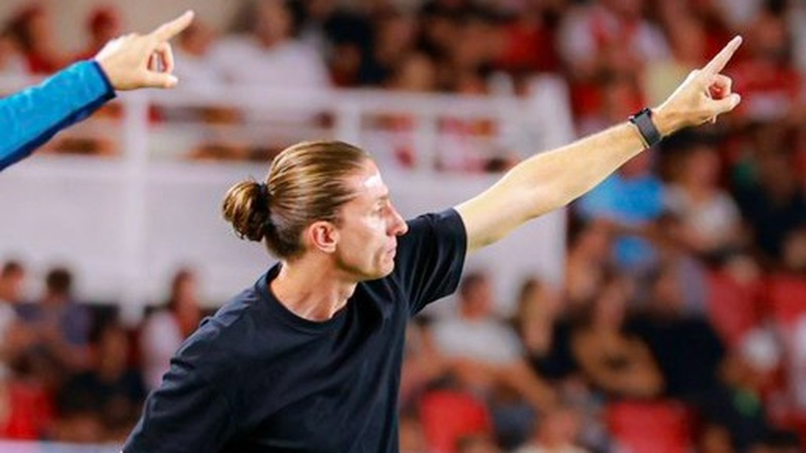 Homem com coque e camiseta preta aponta, em estádio com público ao fundo