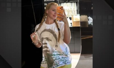 Mulher loira de tranças posa para selfie no espelho, vestindo camiseta branca estampada