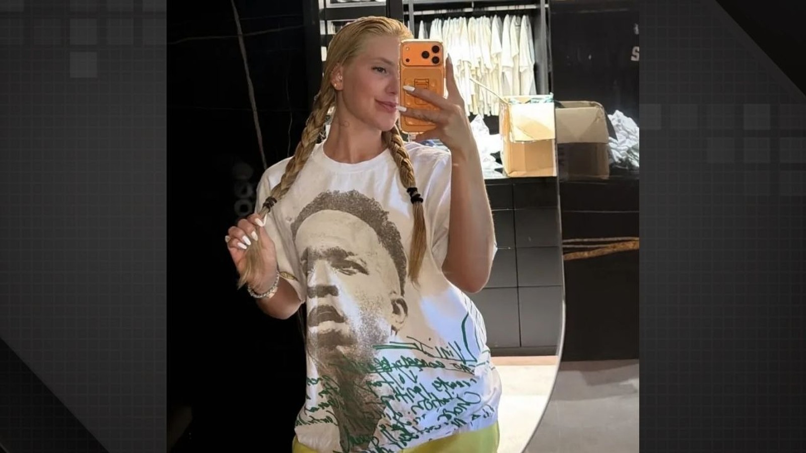 Mulher loira de tranças posa para selfie no espelho, vestindo camiseta branca estampada