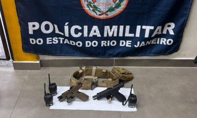 Duas pistolas, dois rádios e cinto tático sobre o chão com banner da Polícia Militar