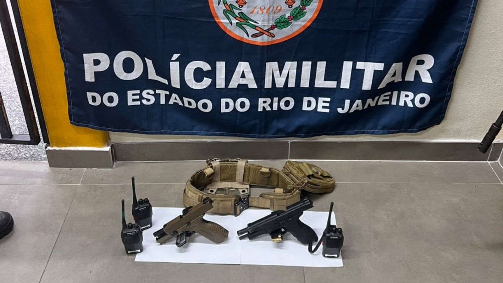 Duas pistolas, dois rádios e cinto tático sobre o chão com banner da Polícia Militar