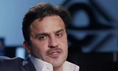 Homem, Pablo Marçal, de terno, camisa branca e bigode, em fundo desfocado azul