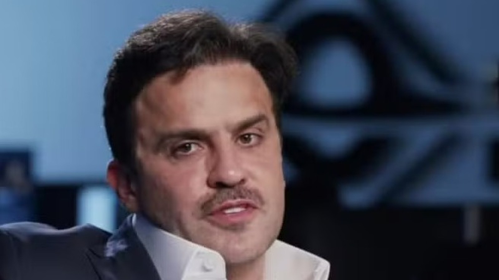 Homem, Pablo Marçal, de terno, camisa branca e bigode, em fundo desfocado azul