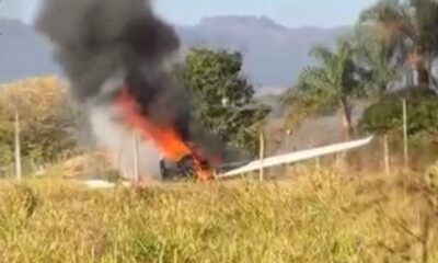 Pequeno avião em chamas com fumaça preta em campo de grama e árvores