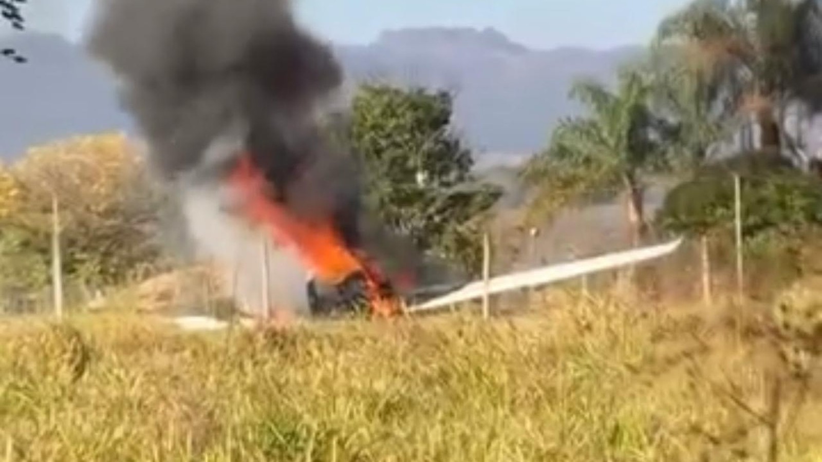 Pequeno avião em chamas com fumaça preta em campo de grama e árvores