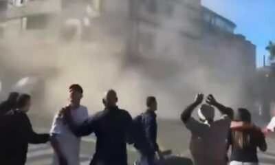 Pessoas em rua empoeirada reagem a evento, com edifícios danificados ao fundo