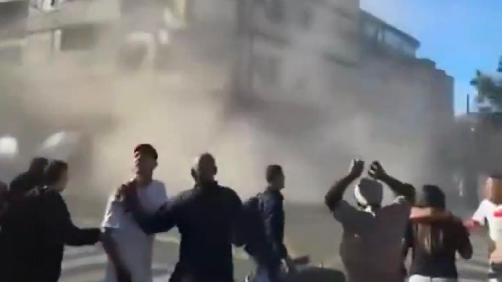 Pessoas em rua empoeirada reagem a evento, com edifícios danificados ao fundo