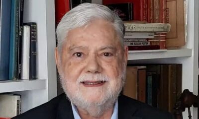 Retrato de Tito Bruno Ryff, homem idoso de cabelo e barba grisalhos, sorrindo, com estante de livros ao fundo