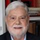 Retrato de Tito Bruno Ryff, homem idoso de cabelo e barba grisalhos, sorrindo, com estante de livros ao fundo