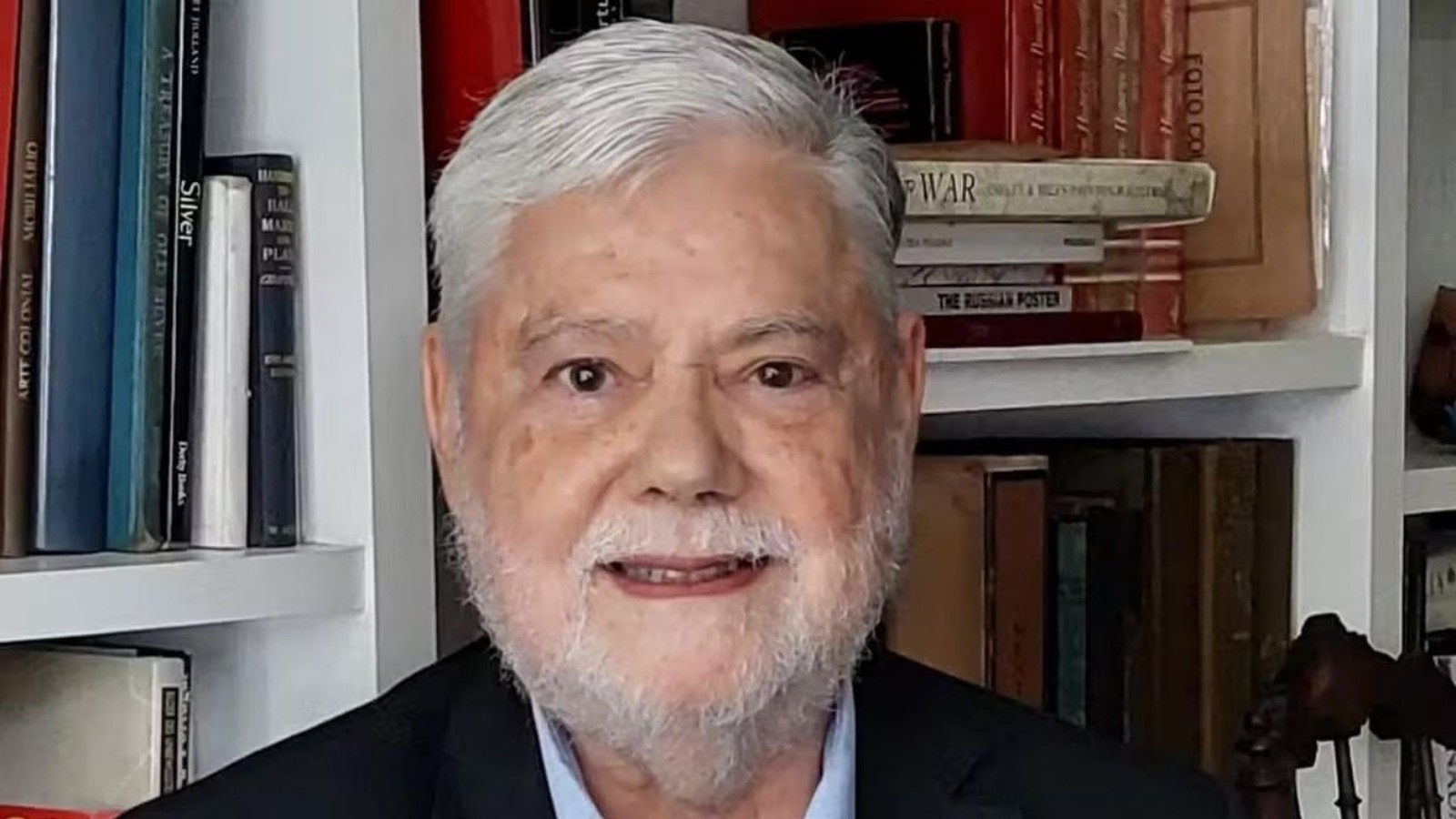 Retrato de Tito Bruno Ryff, homem idoso de cabelo e barba grisalhos, sorrindo, com estante de livros ao fundo