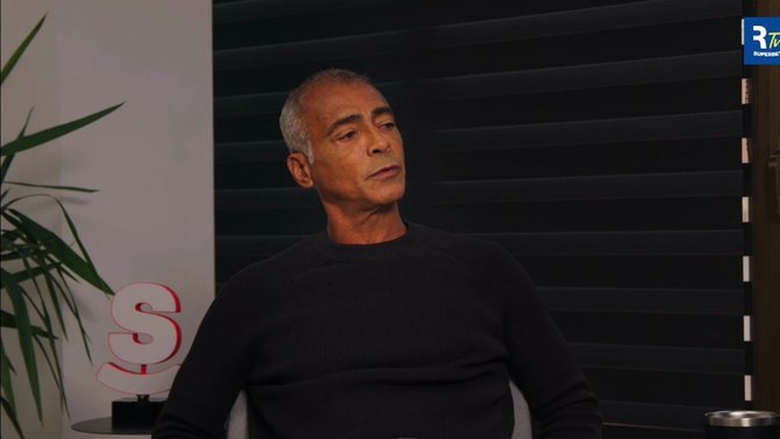 Romário, cabelo grisalho, camisa escura, sentado, olhando para a direita em ambiente interno