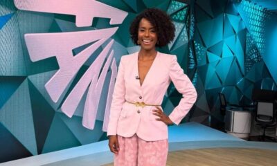 Maju Coutinho sorrindo em estúdio com blazer rosa, cinto dourado e fundo geométrico azul