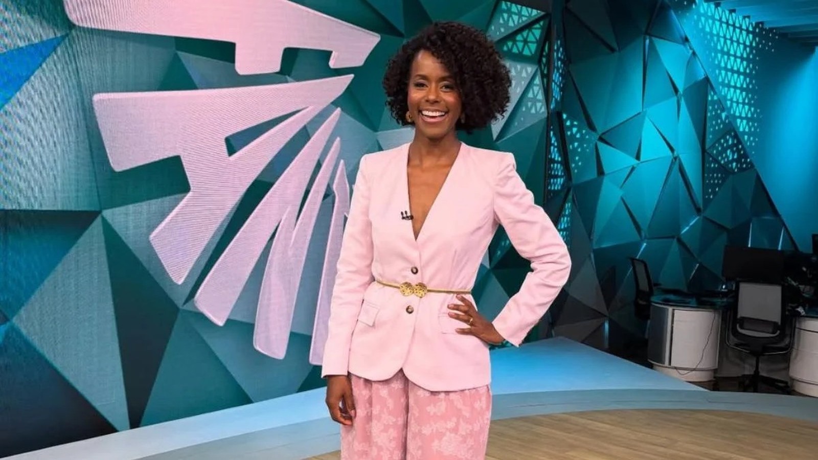 Maju Coutinho sorrindo em estúdio com blazer rosa, cinto dourado e fundo geométrico azul