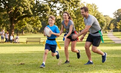 Família jogando tag rugby em um parque, com criança correndo com a bola enquanto os pais participam.